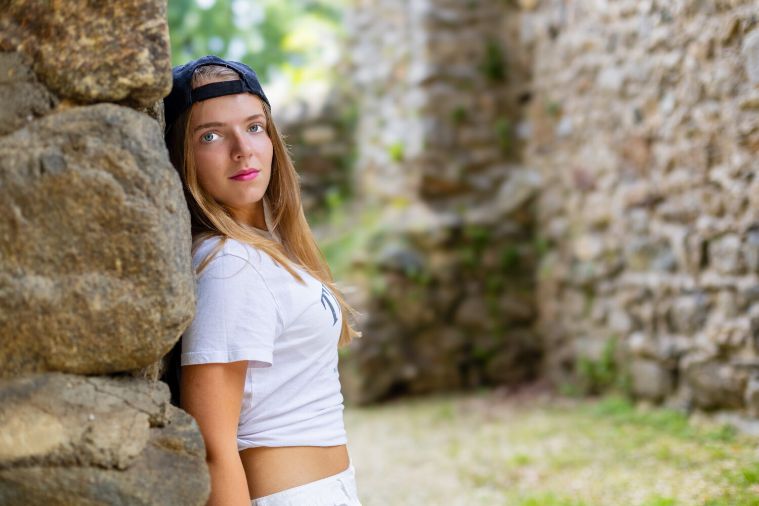 Teenagerin in weißem T-Shirt und Baseballkappe lehnt an einer alten Steinmauer